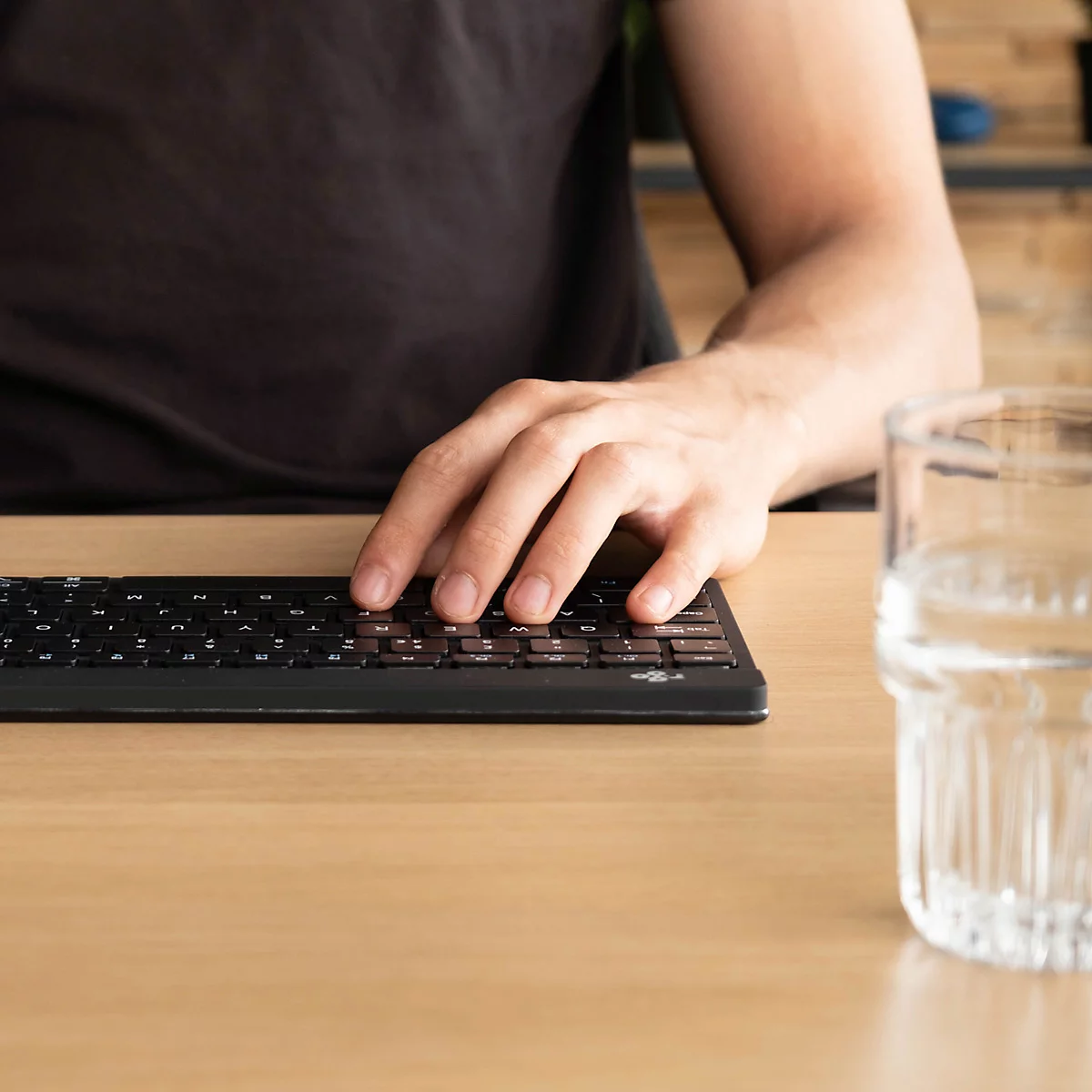 Une personne utilise un clavier noir sur un bureau brun clair. Un verre d'eau est à côté.