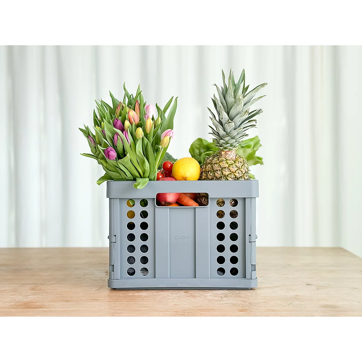 Grey crate with fruits, vegetables and flowers, on light table. Pineapple, lettuce, lemon, carrot and tulips are visible.