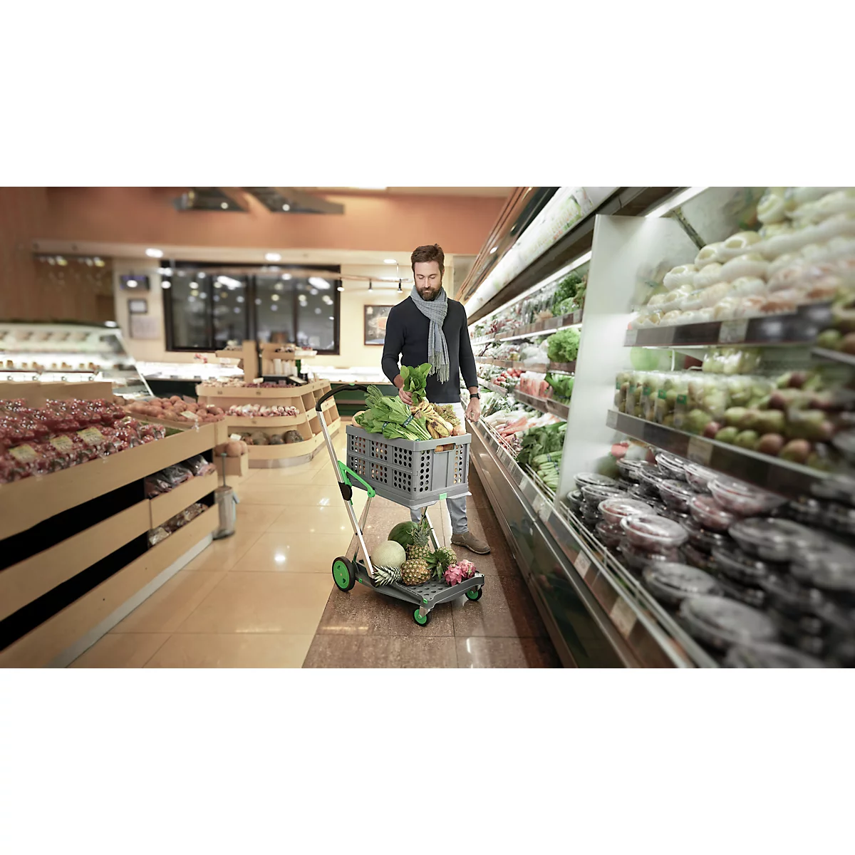 Man in a supermarket with a shopping cart filled with vegetables. He is standing in front of a refrigerated section with vegetables.