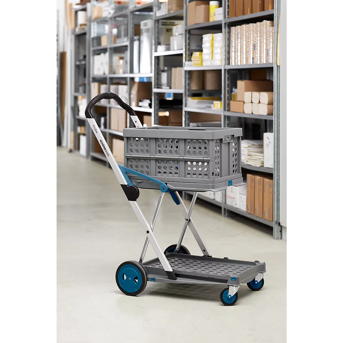 Grauer Klappwagen mit Kiste, in einem Lagerhaus vor Regalen mit Kartons. Blaue Räder und Griffe.