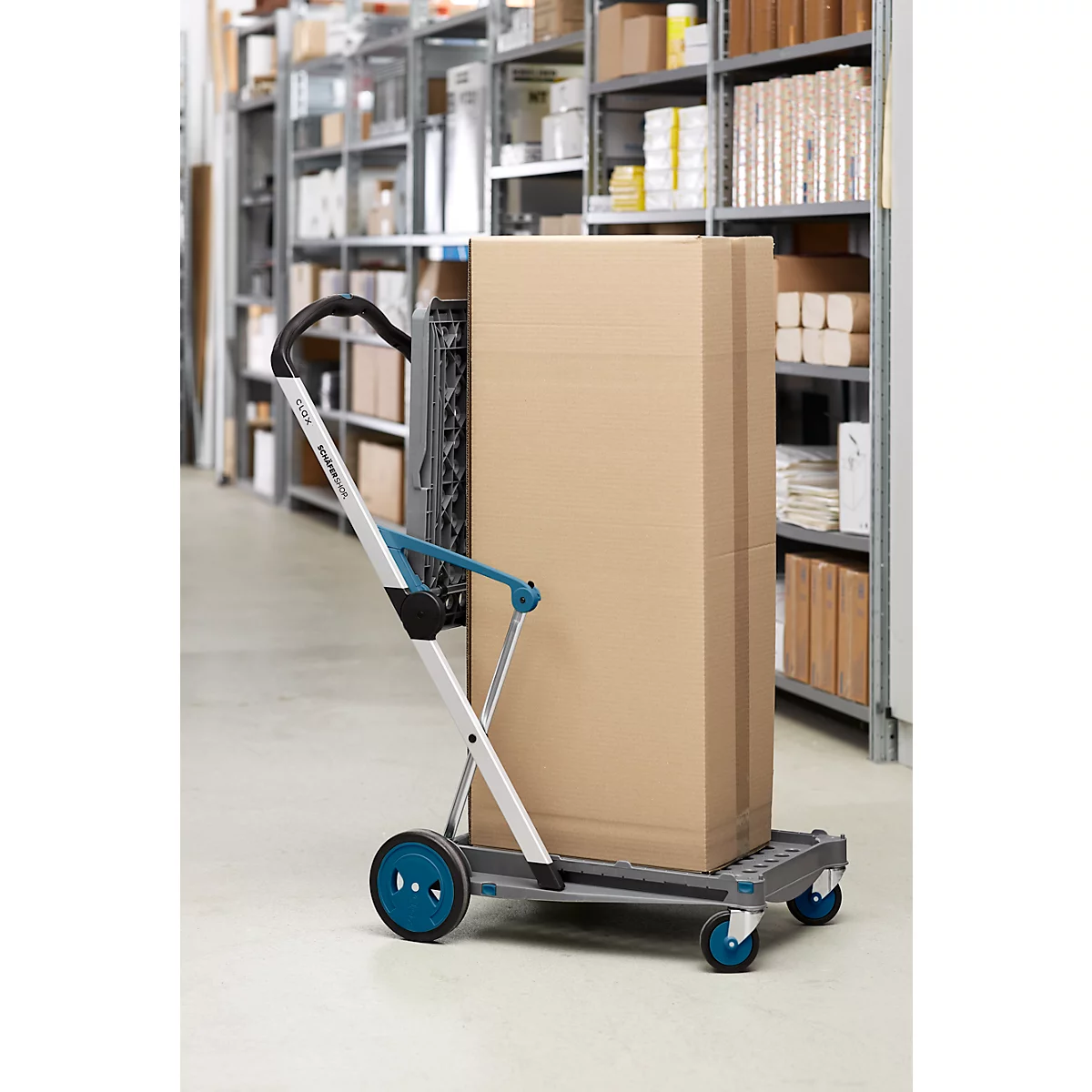 Ein grauer Transportwagen mit blaugrünen Rädern und Griffen transportiert einen grossen Karton in einem Lagerraum.