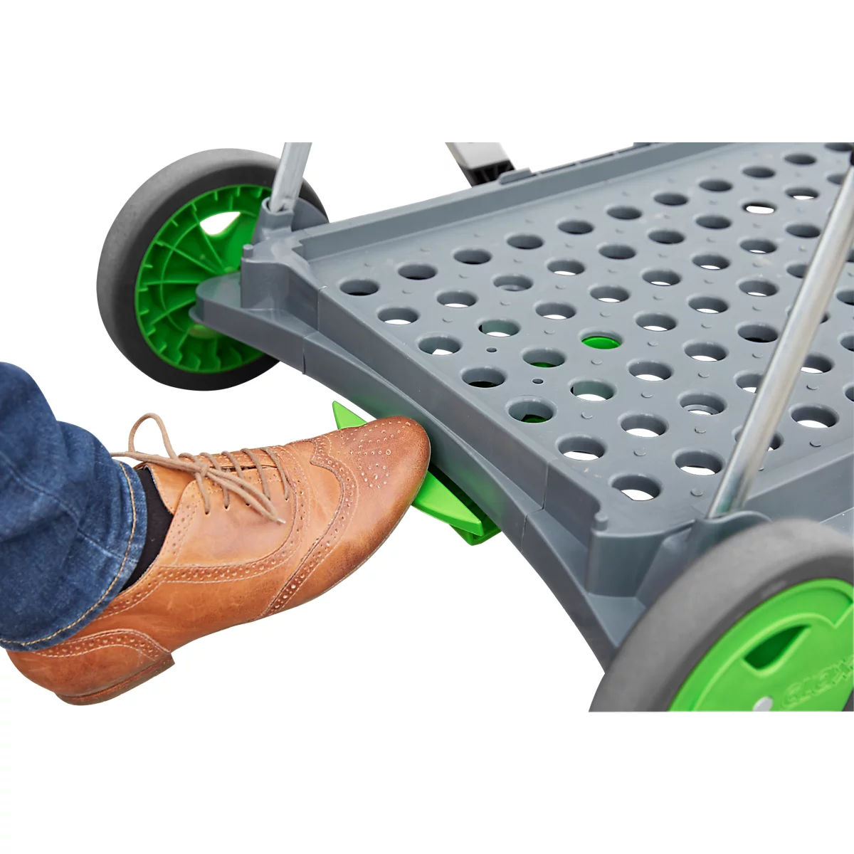 Primer plano de un zapato marrón pisando un pedal verde en un carro gris plegable con ruedas verdes.