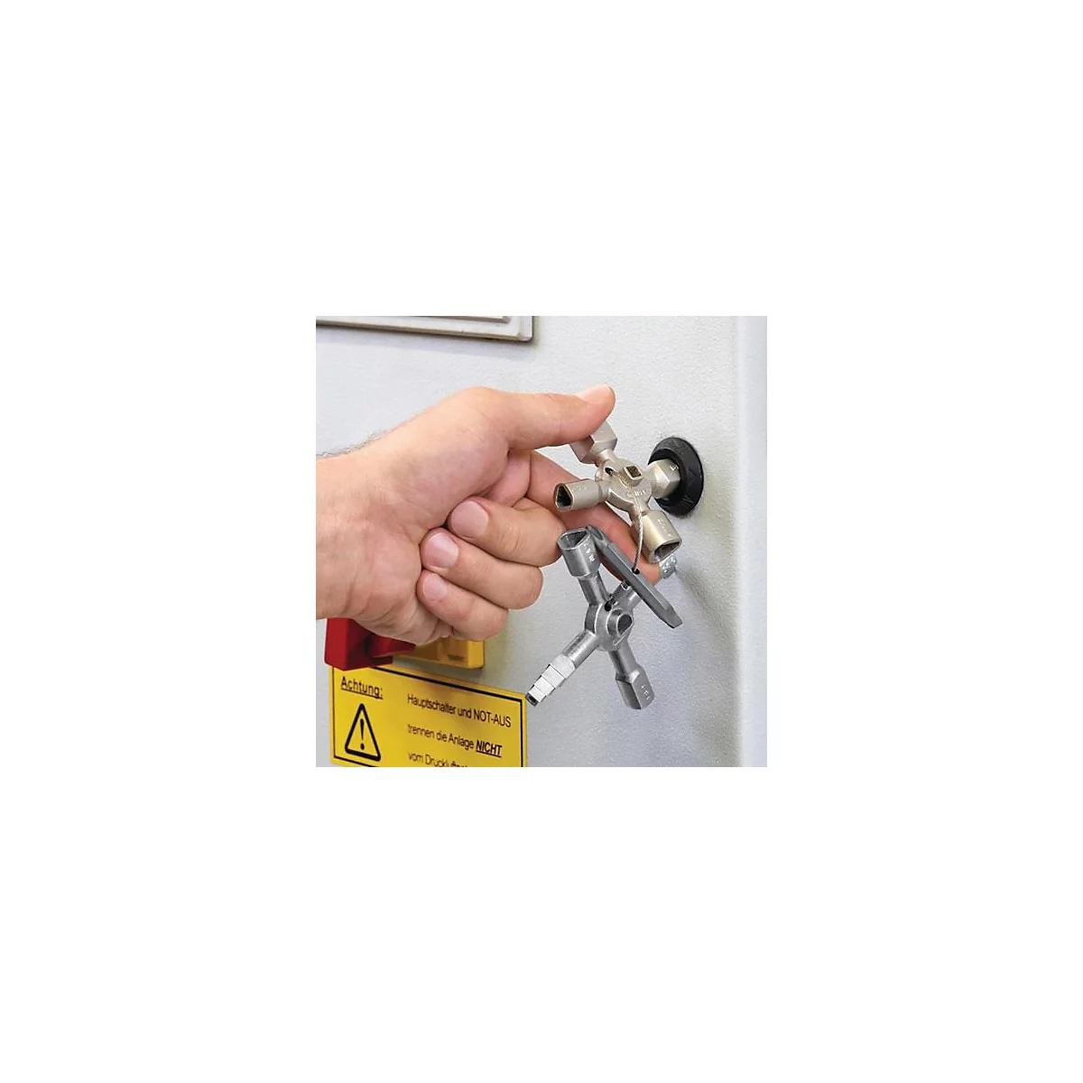Main tenant une clé multifonction sur fond blanc. Sur un panneau, il est écrit : Attention ! L'interrupteur principal et l'arrêt d'urgence ne déconnectent pas l'installation.