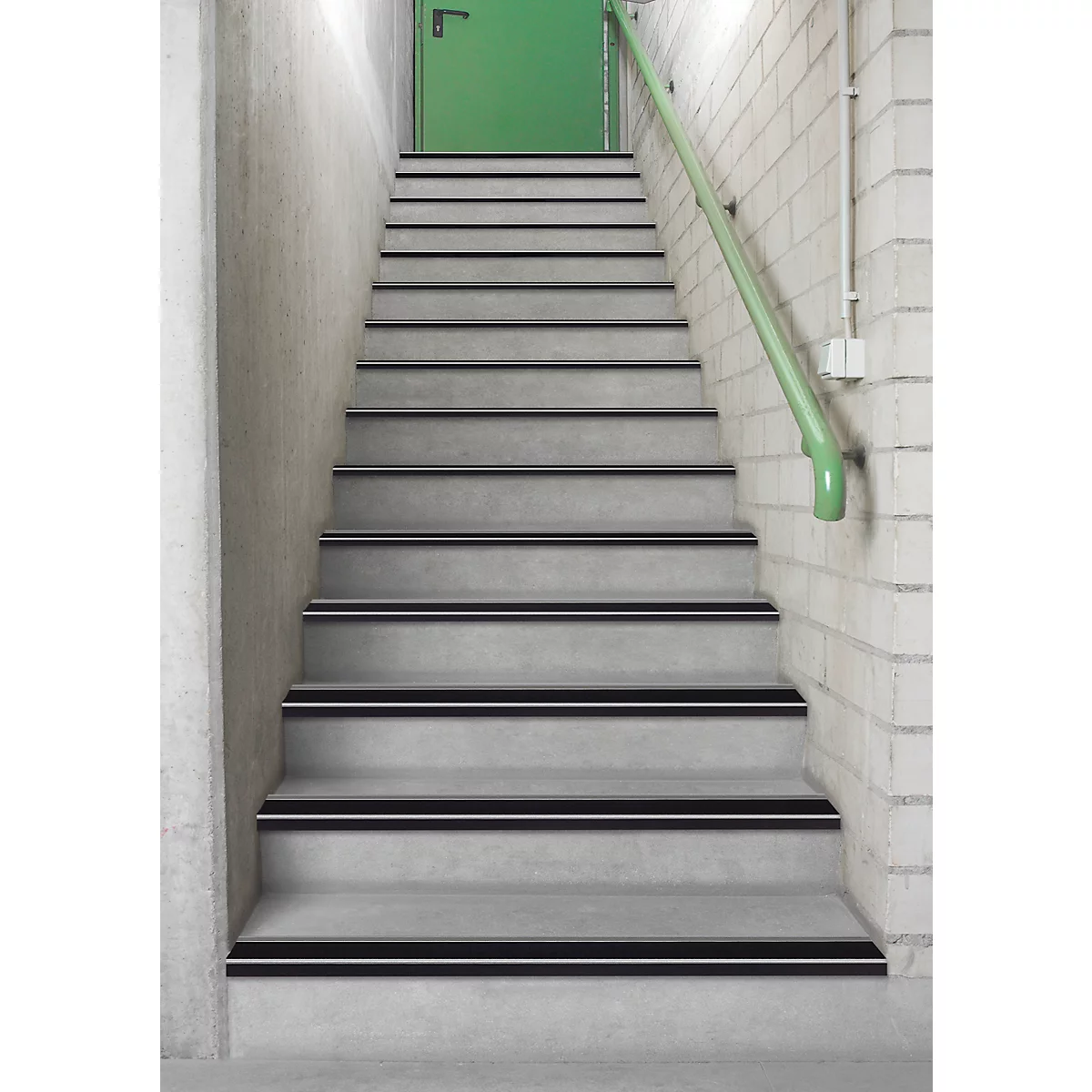 Stairs with gray steps, black stripes and a green handrail. At the end of the stairs is a green door.