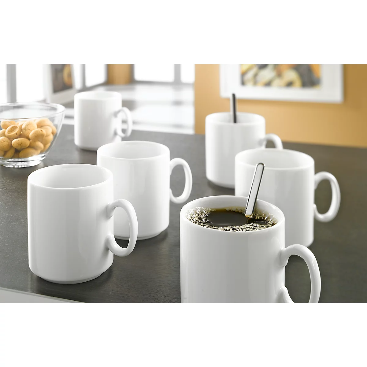 White coffee cups on a table. One cup with coffee, one with a spoon, nuts in a glass bowl.