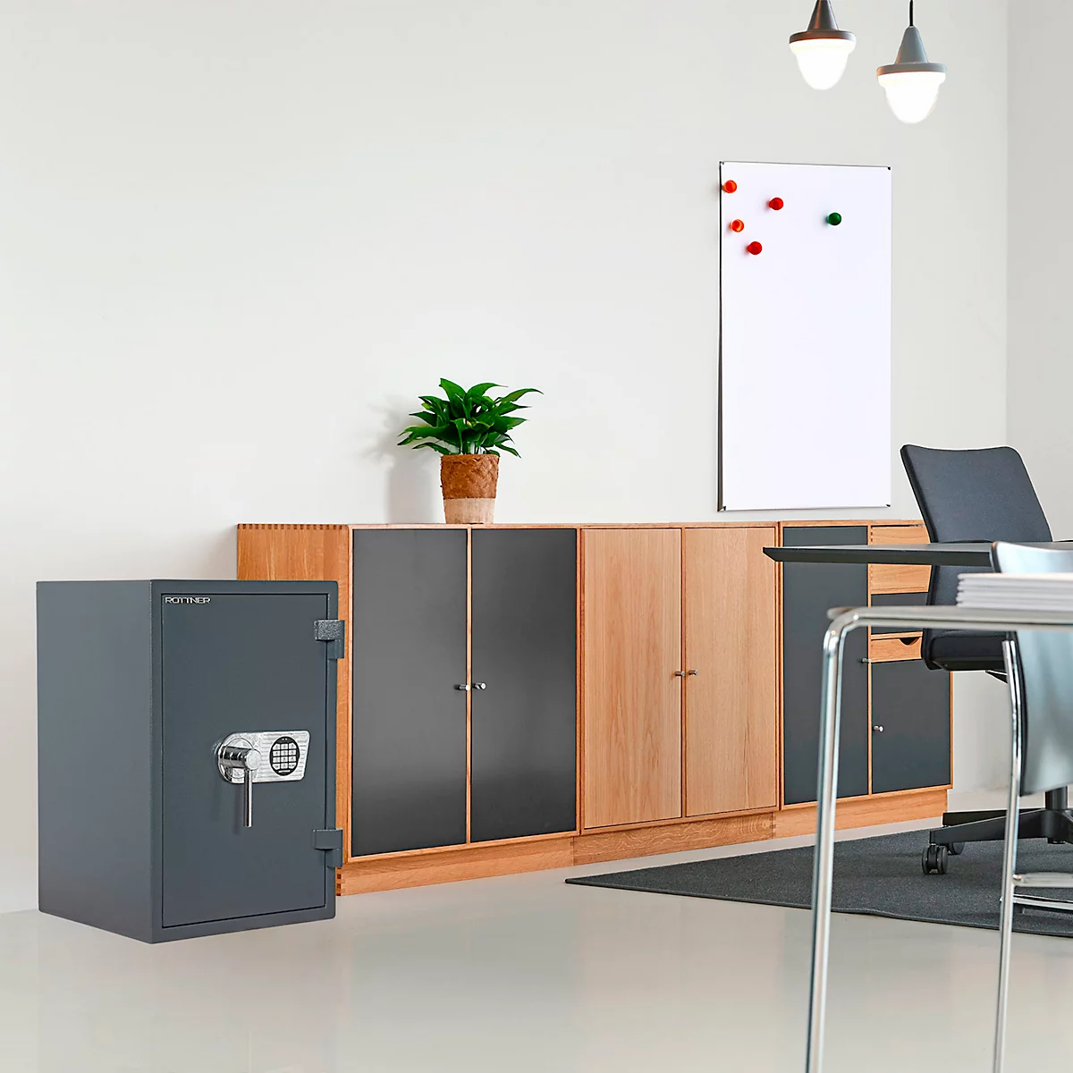 Coffre-fort gris devant des meubles de bureau, des plantes et un tableau blanc. Le coffre-fort est équipé d'un pavé numérique.