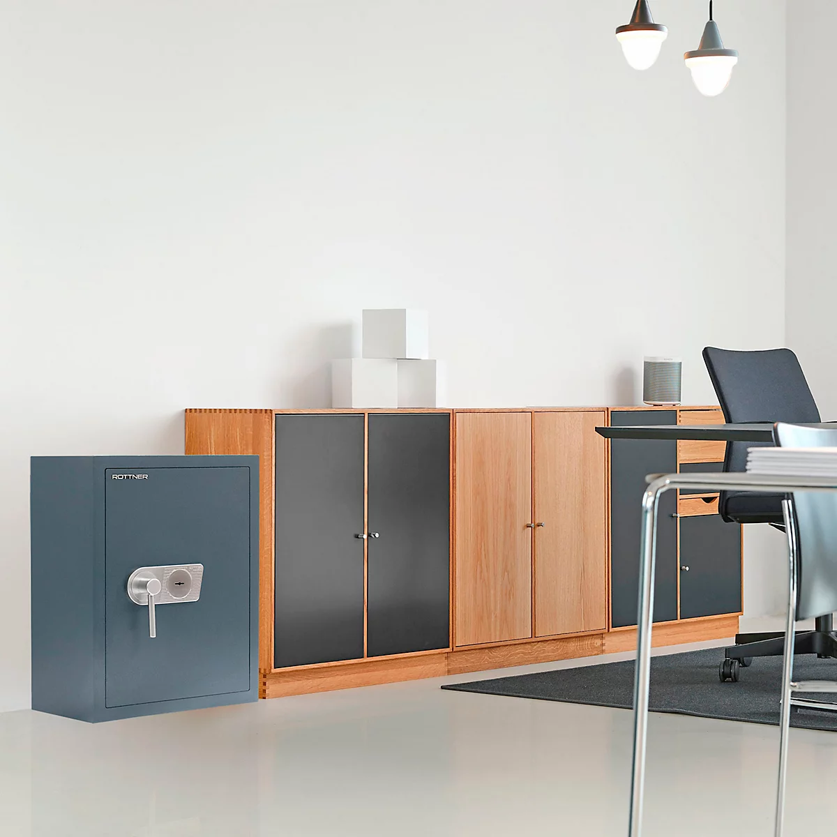 Coffre-fort bleu-gris dans un bureau lumineux. Derrière, des armoires en bois, une table et une chaise.