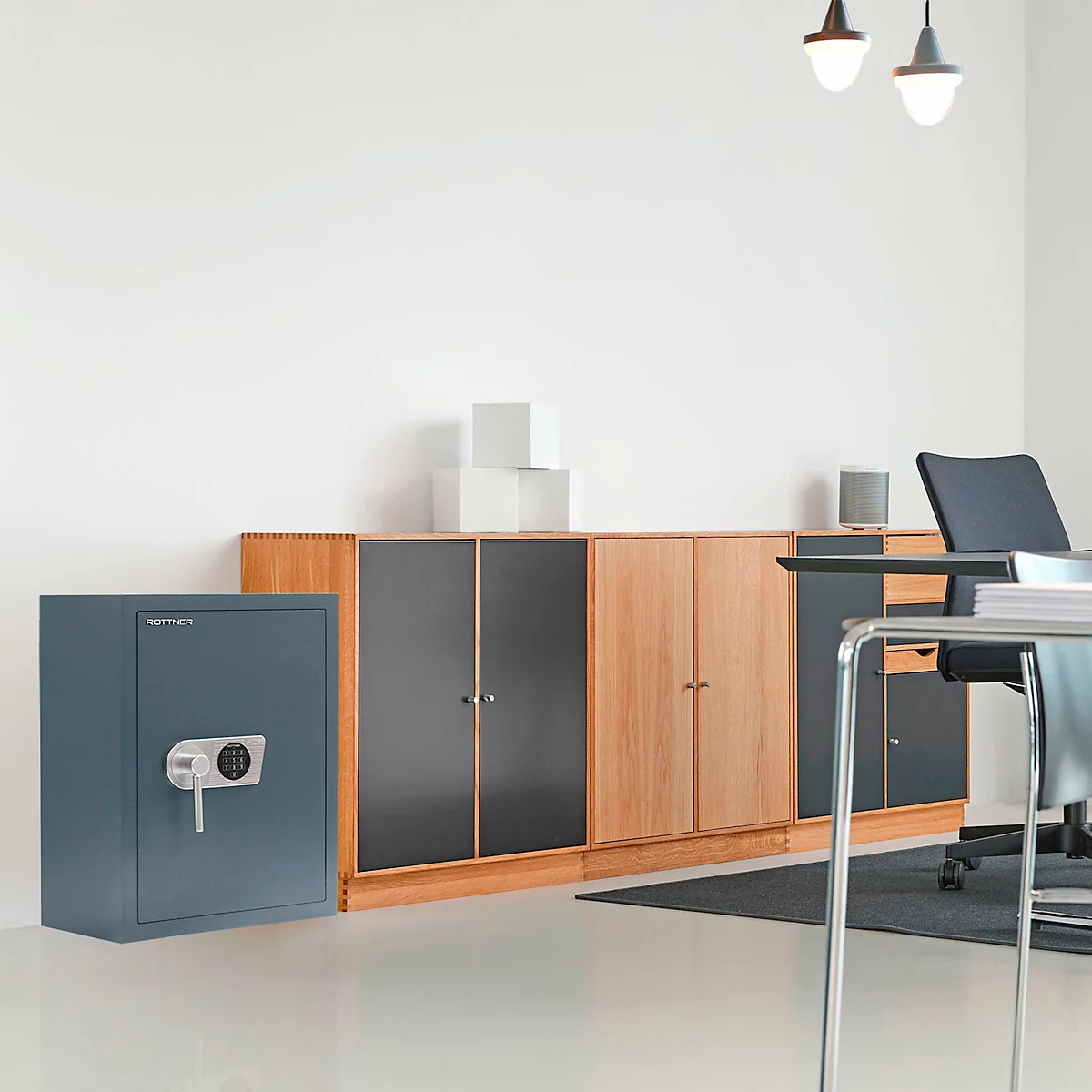 Un coffre-fort bleu est situé à gauche à côté d'une armoire en bois dans un bureau. En arrière-plan : bureau, chaises, lampes.