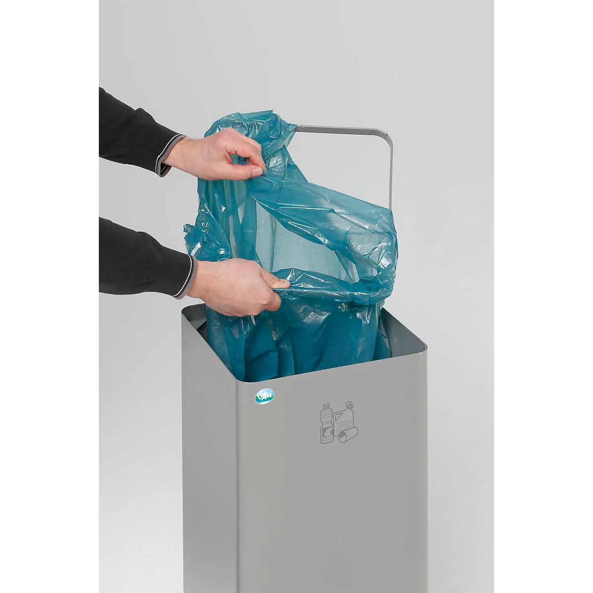 Des mains placent un sac poubelle bleu dans une poubelle grise.