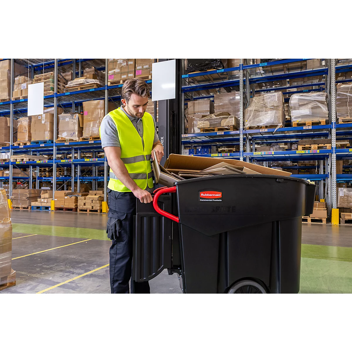 Un homme dans un entrepôt avec un gilet de sécurité et un chariot à ordures noir. Il y met du carton.