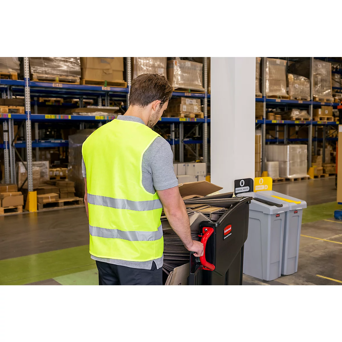 Un homme en gilet de sécurité pousse un chariot de déchets noir dans un entrepôt. Des étagères avec des marchandises sont visibles en arrière-plan.