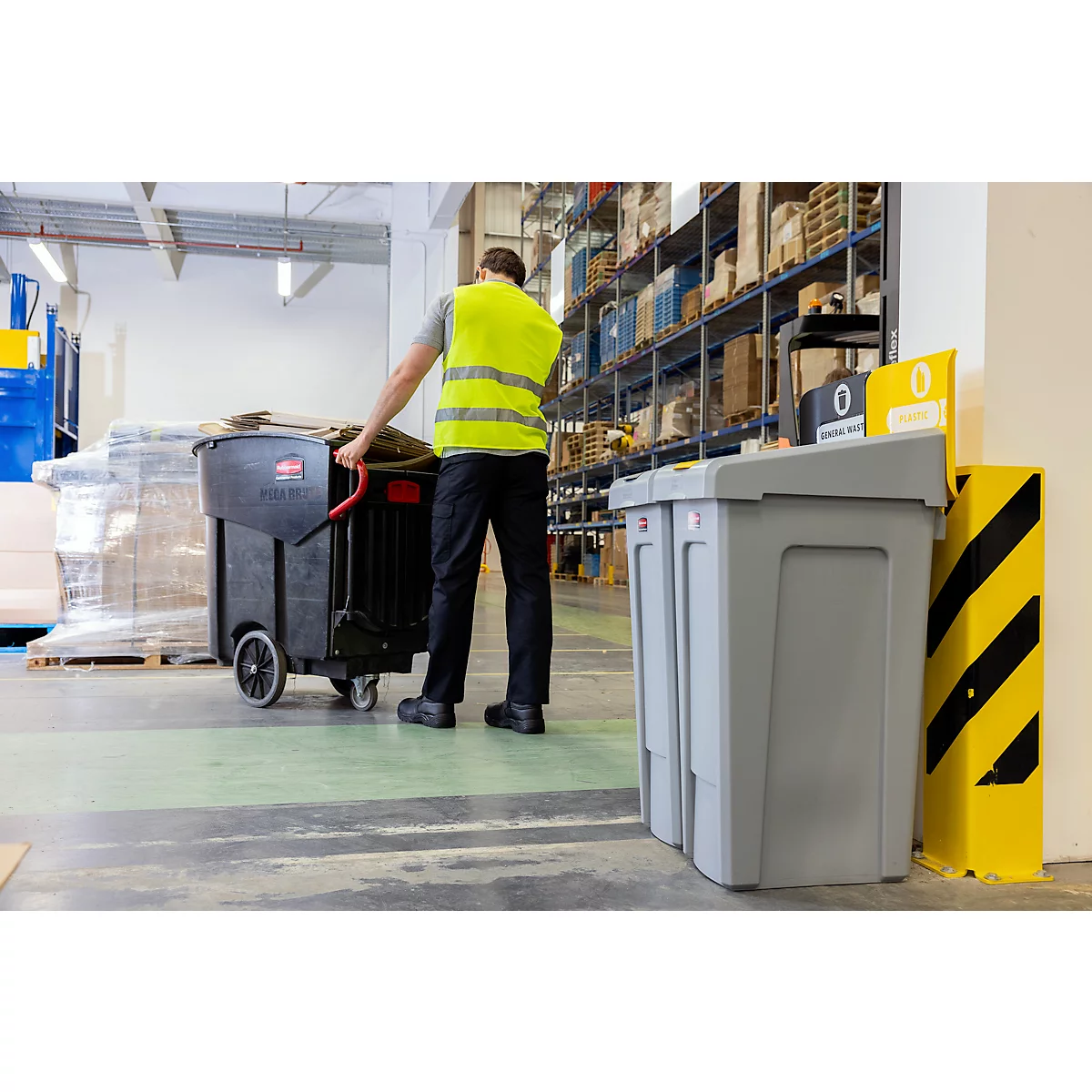 Un employé tire un chariot à déchets noir dans un entrepôt. On aperçoit des étagères et un poteau gris et jaune en arrière-plan.