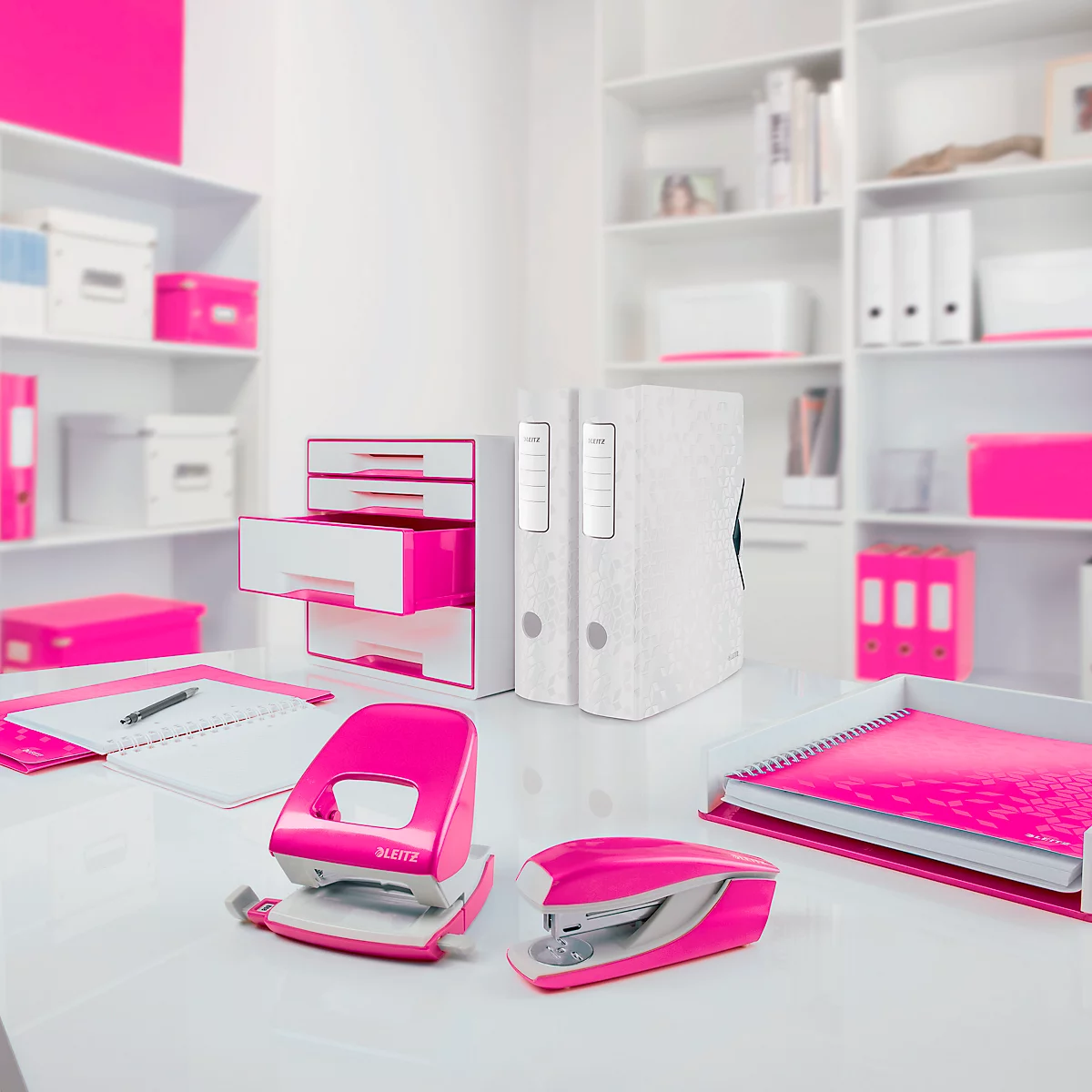 Ein Schreibtisch mit pinkfarbenen Büroartikeln: Locher, Hefter und Notizbücher. Regale im Hintergrund.