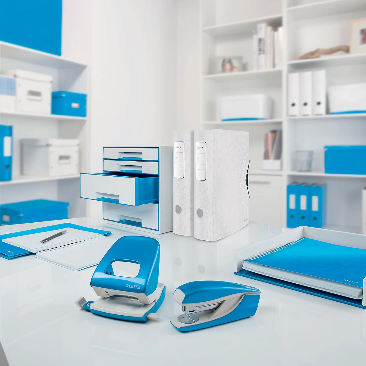 Büro mit weißem Schreibtisch. Locher und Tacker in Blau. Ordner, Schubladen und Boxen in Weiß und Blau.