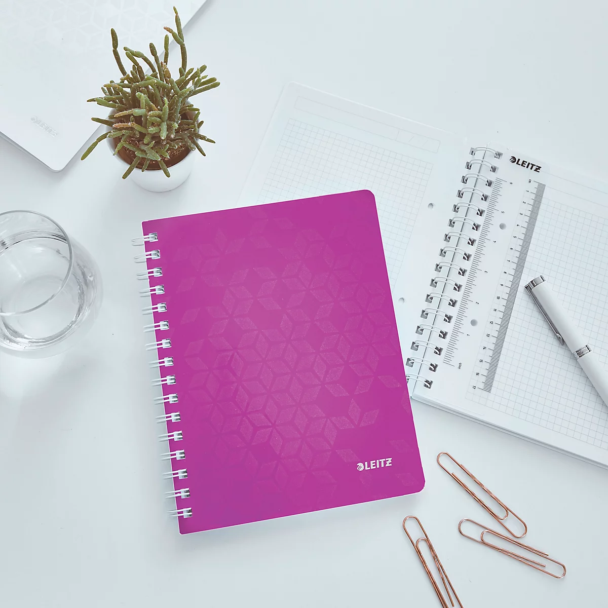 Ein pinkfarbenes Notizbuch mit Spiralbindung liegt auf einem weißen Schreibtisch. Daneben: ein Lineal, ein Stift, Büroklammern und eine kleine Pflanze.