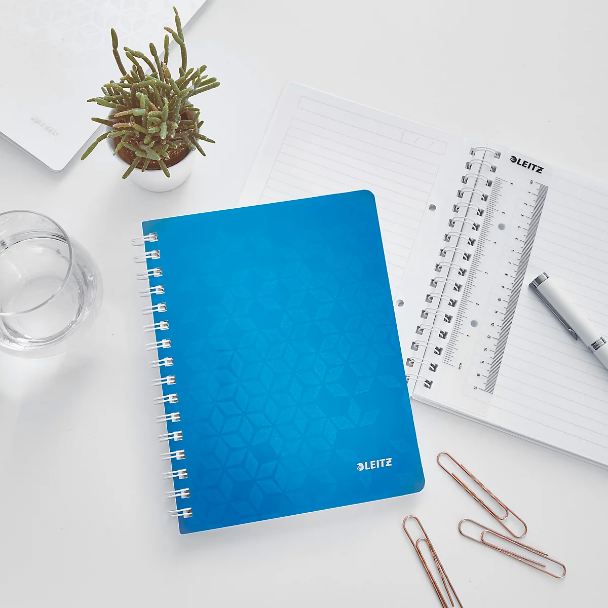 Blaues Notizbuch mit Spirale und Stifthalter, daneben Papier, Stift, Büroklammern und Pflanze auf weißem Tisch.