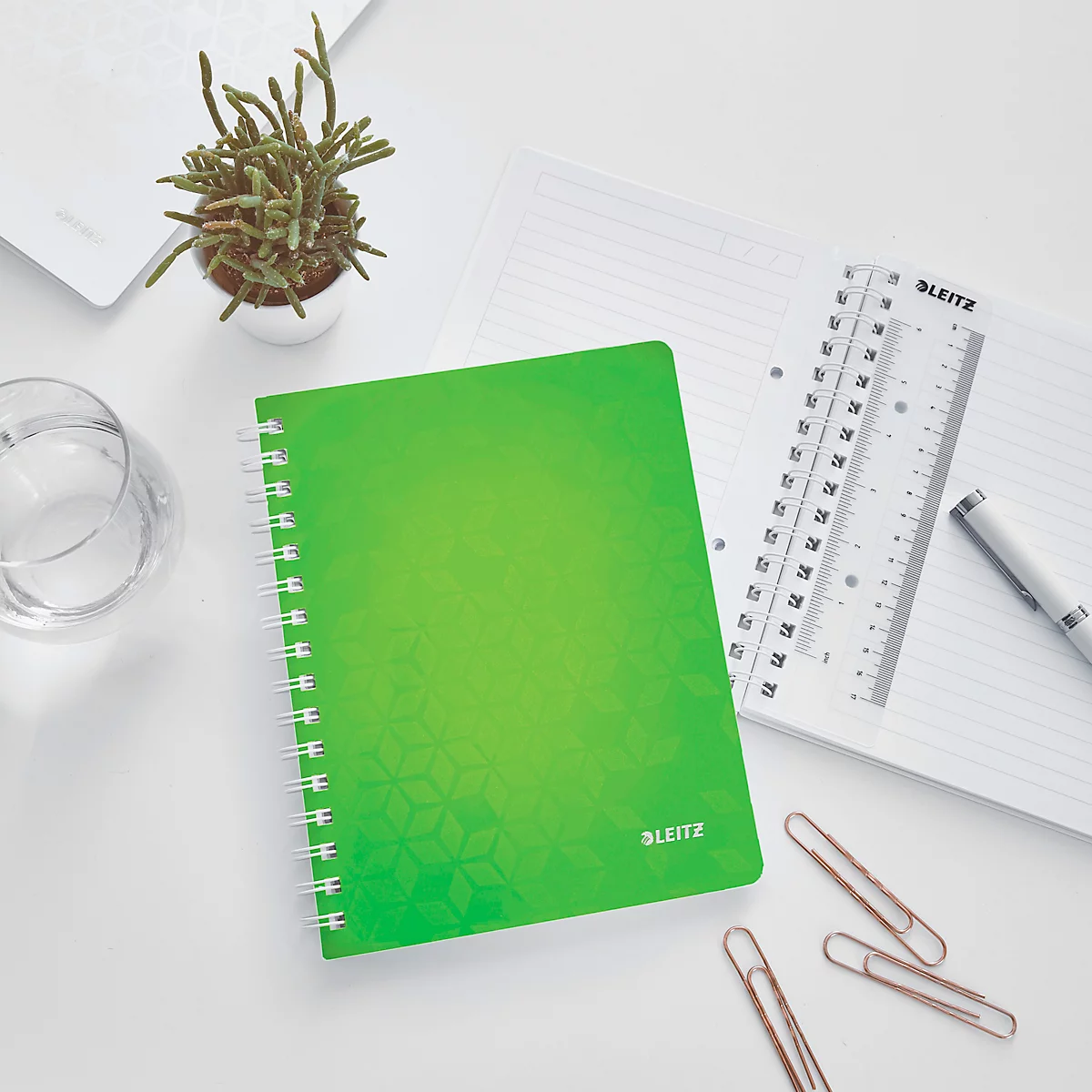 Grünes Notizbuch auf Schreibtisch, neben Stift, Lineal, Büroklammern und Pflanze.