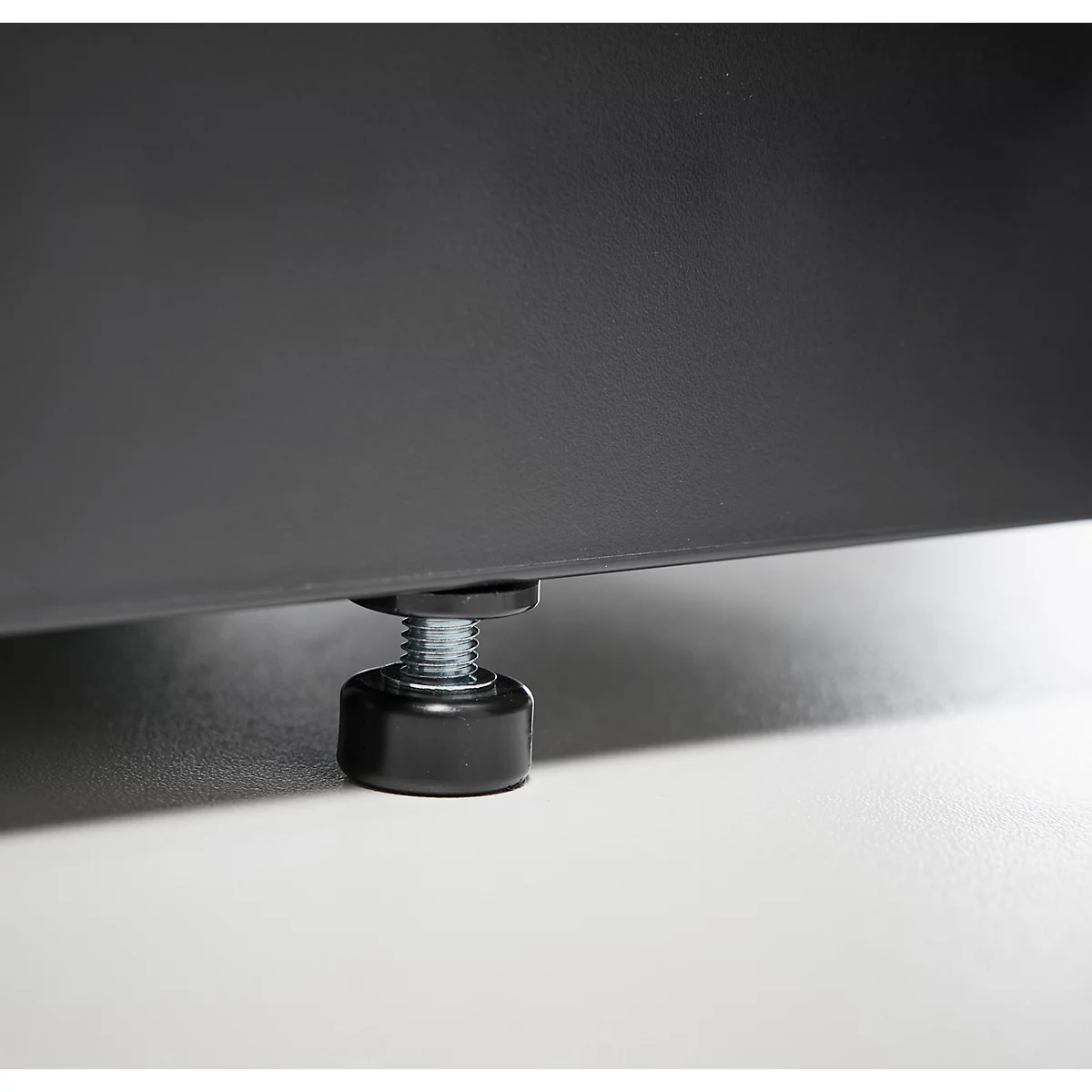 Gros plan d'un pied de meuble réglable. Un pied noir, cylindrique, avec un boulon fileté argenté, situé sous un panneau sombre, repose sur une surface blanche.