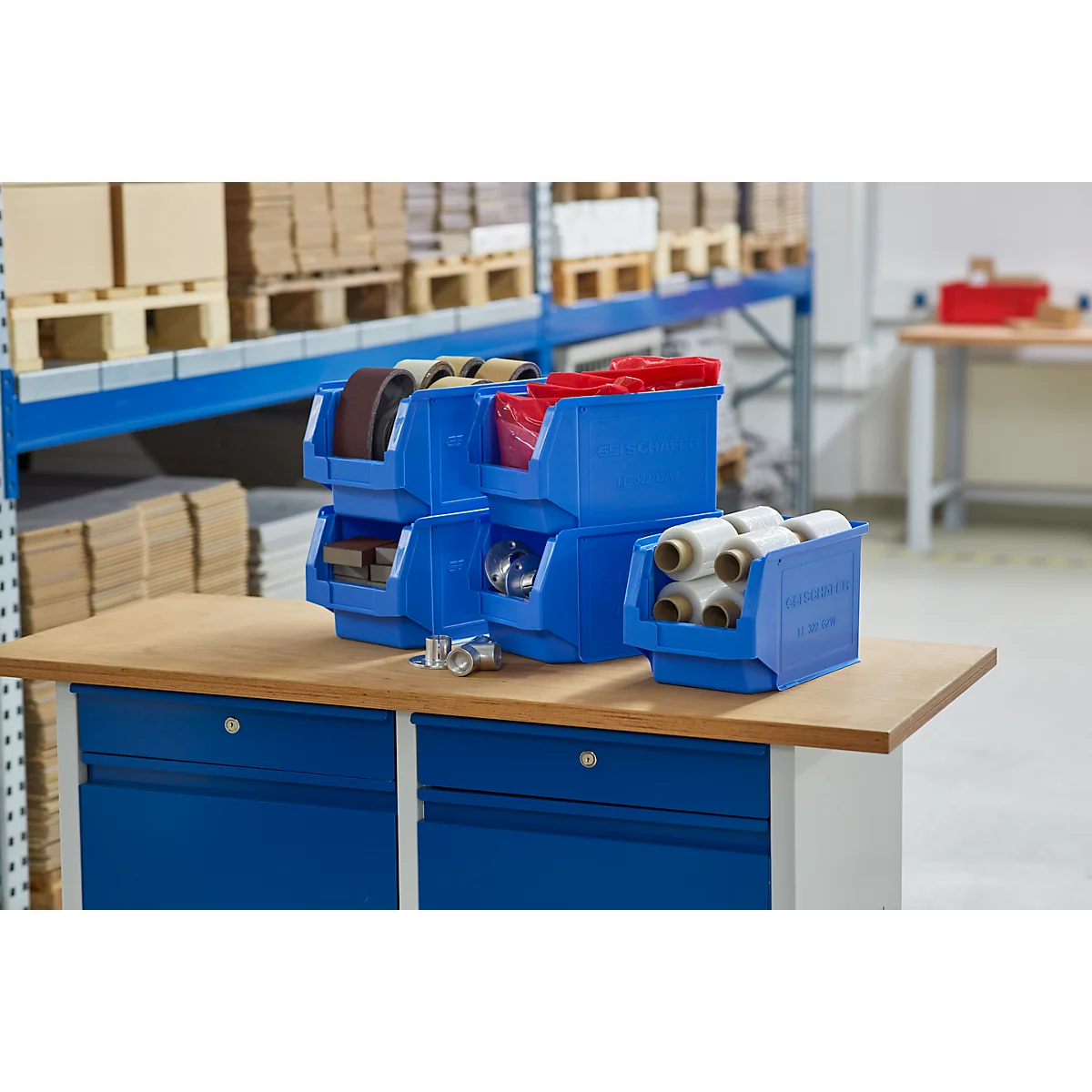 Contenedores azules con varios materiales sobre una mesa de madera frente a una estantería. Los contenedores están llenos de rollos, cintas y otros artículos.