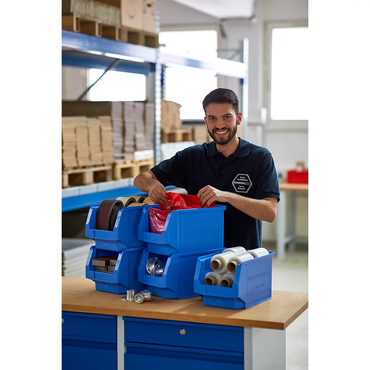Un hombre con ropa de trabajo azul está de pie sonriendo frente a contenedores azules. Sostiene una bolsa roja en la mano.