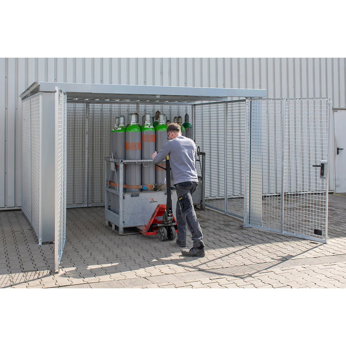 Un homme pousse des bouteilles de gaz sur un transpalette dans une enceinte de sécurité métallique. Enceinte avec porte grillagée ouverte.