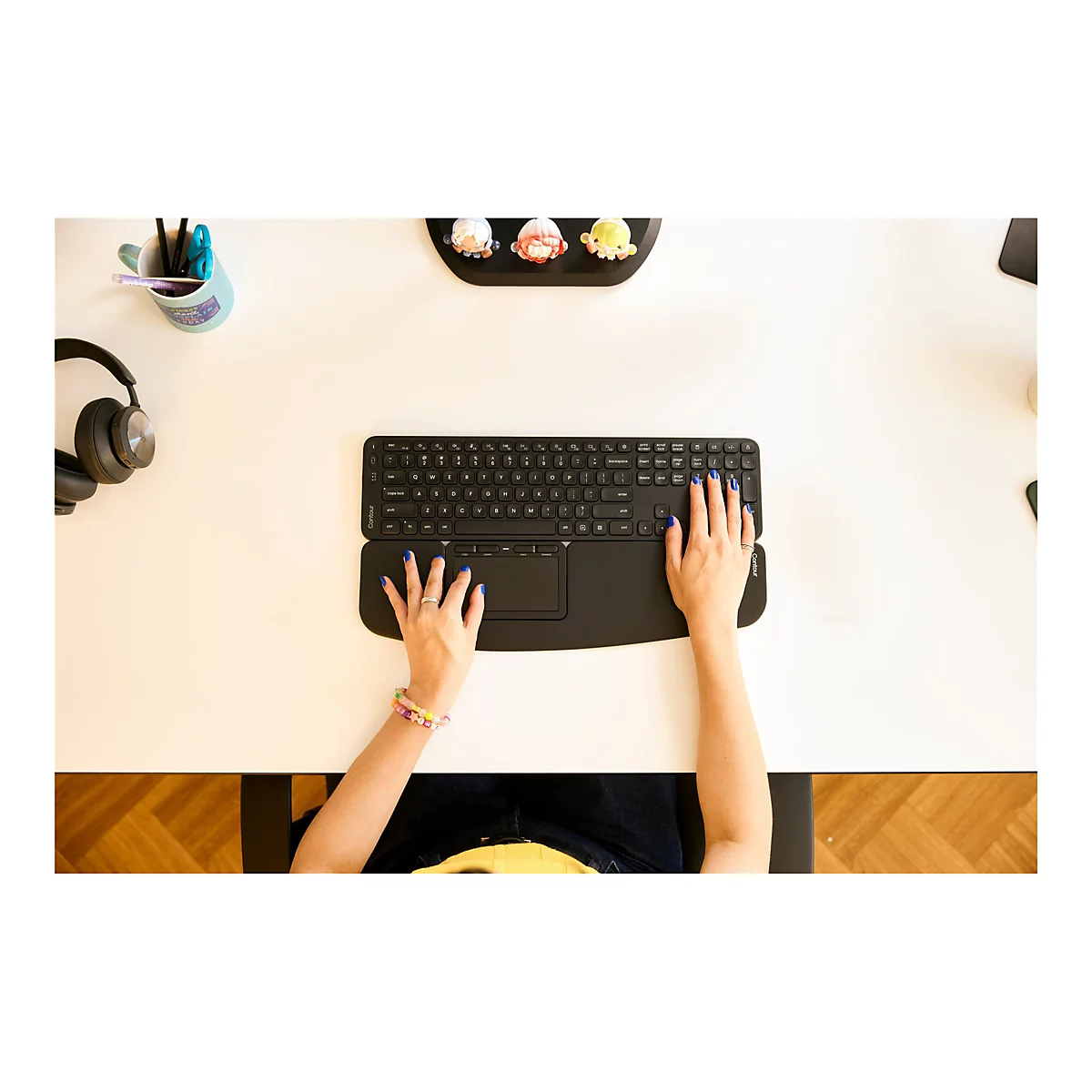 Draufsicht auf einen Schreibtisch mit schwarzer Tastatur und den Händen einer Person. Die Person hat blaue Fingernägel und trägt Ringe und Armbänder.