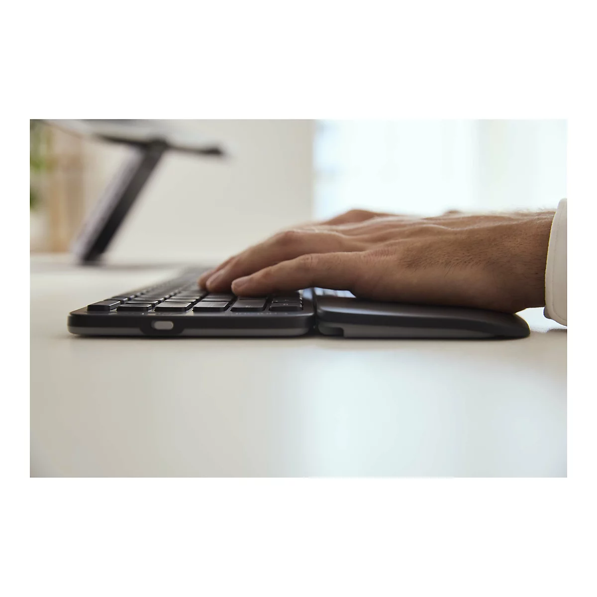 Nahaufnahme einer Person, die eine flache, ergonomische Tastatur bedient. Die Hand liegt auf der Tastatur und einem Handgelenkstütz. Der Hintergrund ist unscharf.