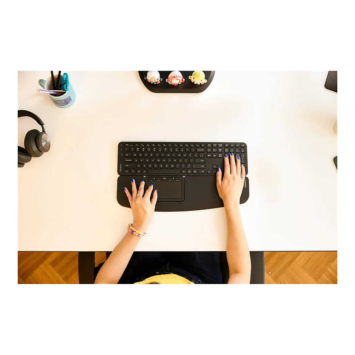Ansicht von oben auf eine Person, die eine schwarze ergonomische Tastatur mit integriertem Trackpad benutzt, blaue Fingernägel. Auf dem Tisch stehen Stifte, eine Tasse und ein Kopfhörer.