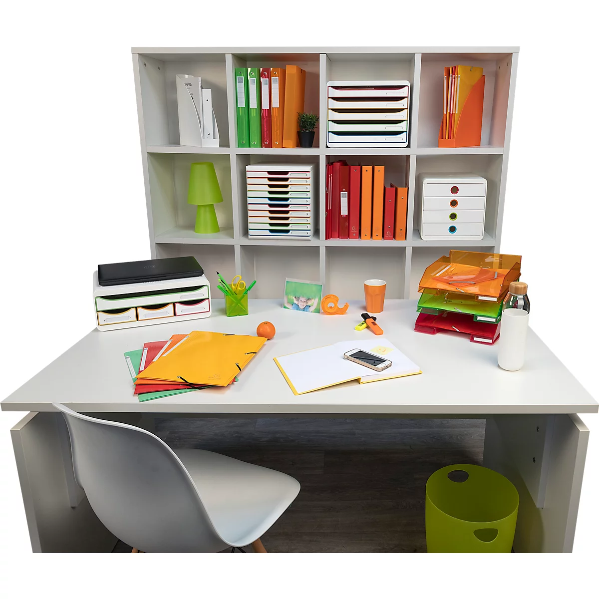 Bureau avec fournitures de bureau : dossiers, plateaux, lampe, tasse et téléphone. Une chaise devant, une étagère derrière.