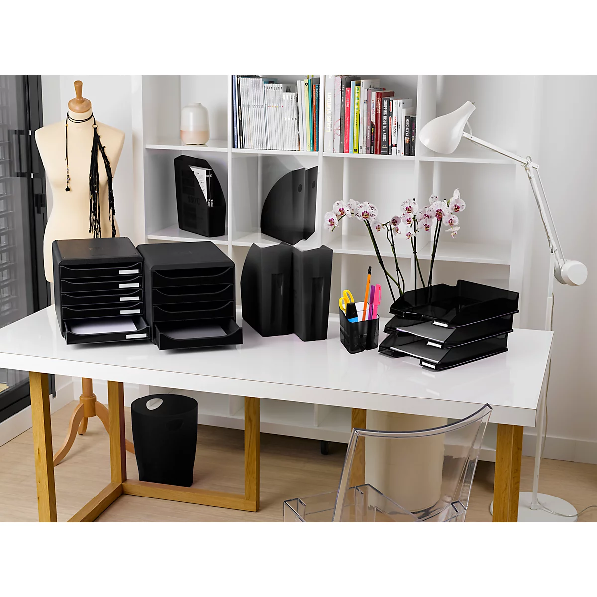 Bureau de bureau avec plateaux noirs, porte-lettres, porte-stylo, lampe, buste de couturière, livres et orchidées.