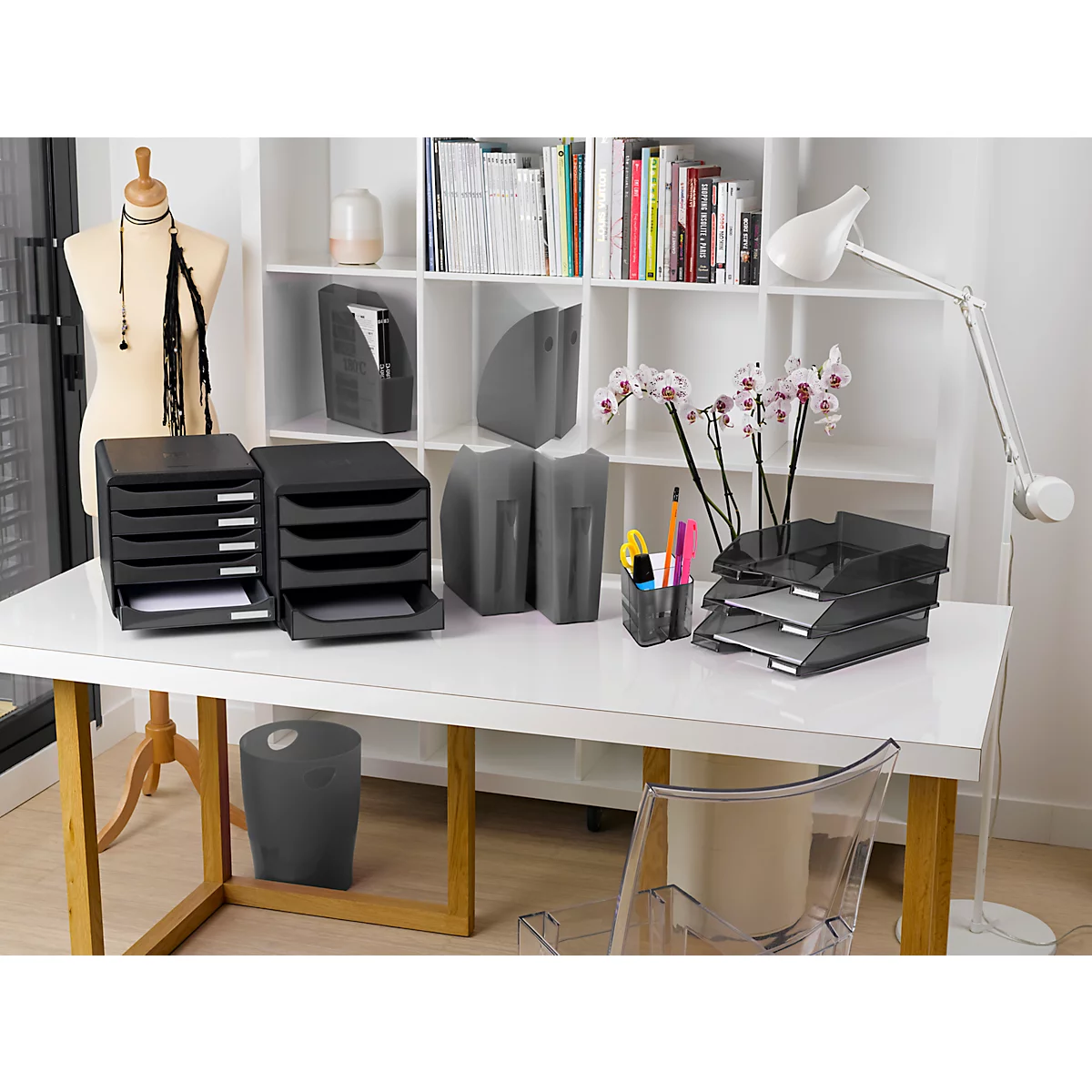 Bureau avec articles d'organisation de bureau : plateaux à papier, porte-stylos, corbeille. Table blanche, bibliothèque derrière.