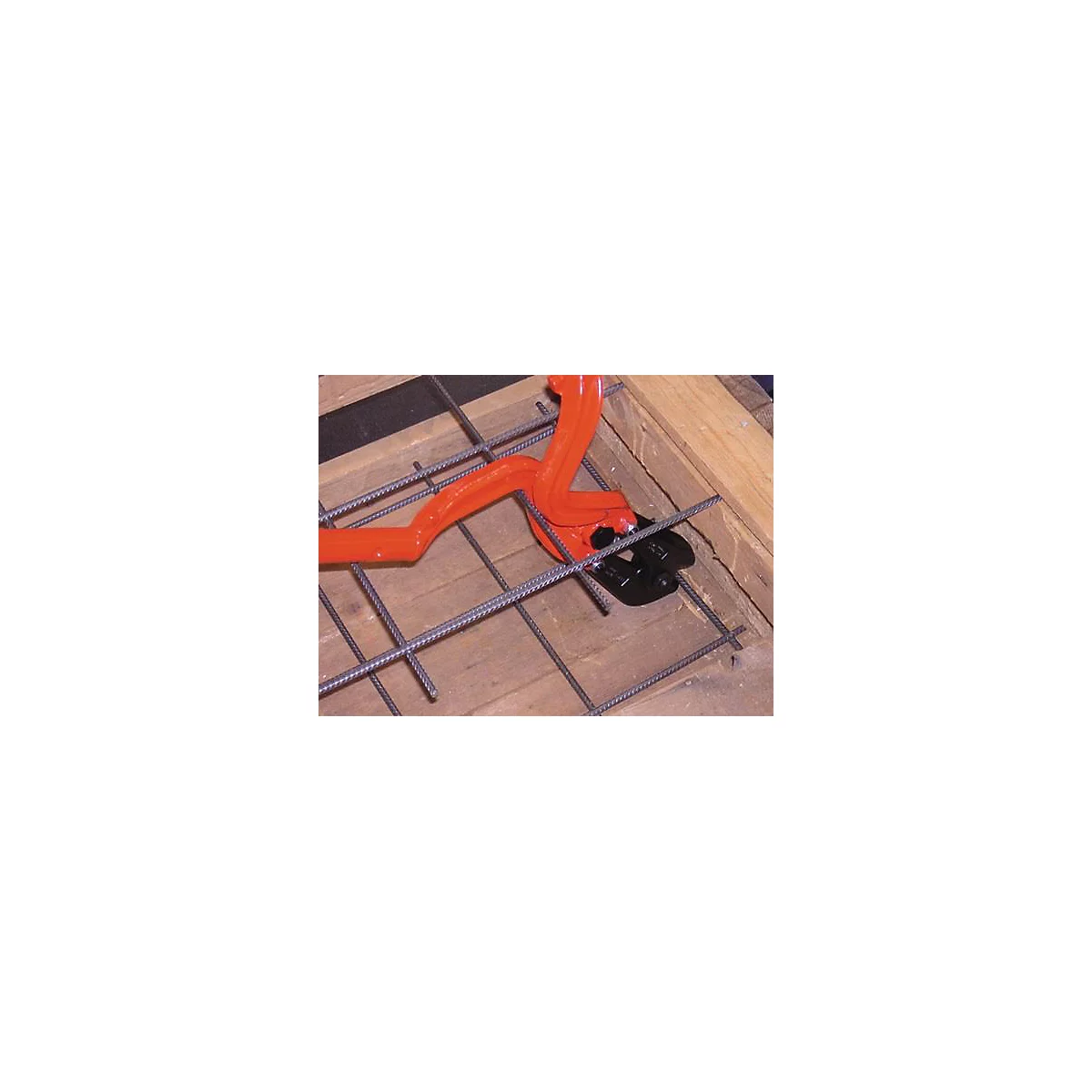 An orange rebar cutter cuts through steel rods lying on a wooden frame.