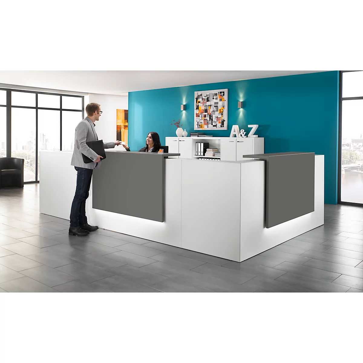 Man at reception desk handing documents. A woman is sitting behind the counter. Behind her blue wall and art.