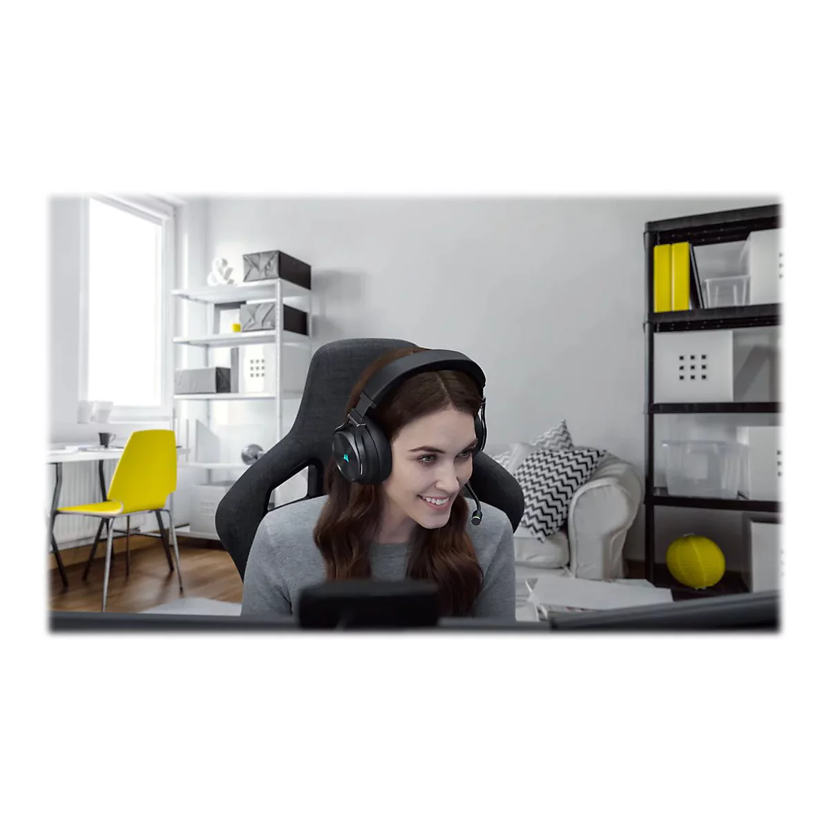Eine lächelnde Frau mit Headset sitzt vor einem Computer. Im Hintergrund: Regal, gelber Stuhl, Sofa.