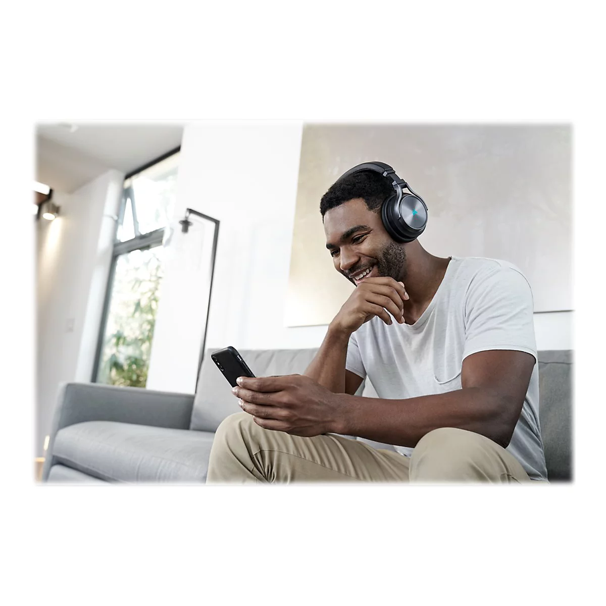 Ein Mann mit Kopfhörern sitzt auf einem Sofa, lächelt und schaut auf sein Smartphone.