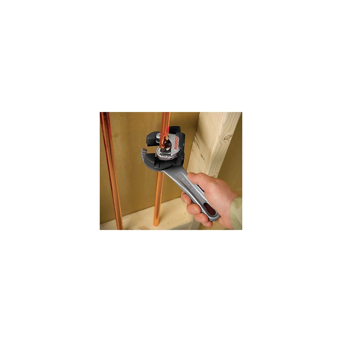 Une main utilise un coupe-tube pour couper un tuyau en cuivre. Le coupe-tube a une poignée argentée et le logo "RIDGID".