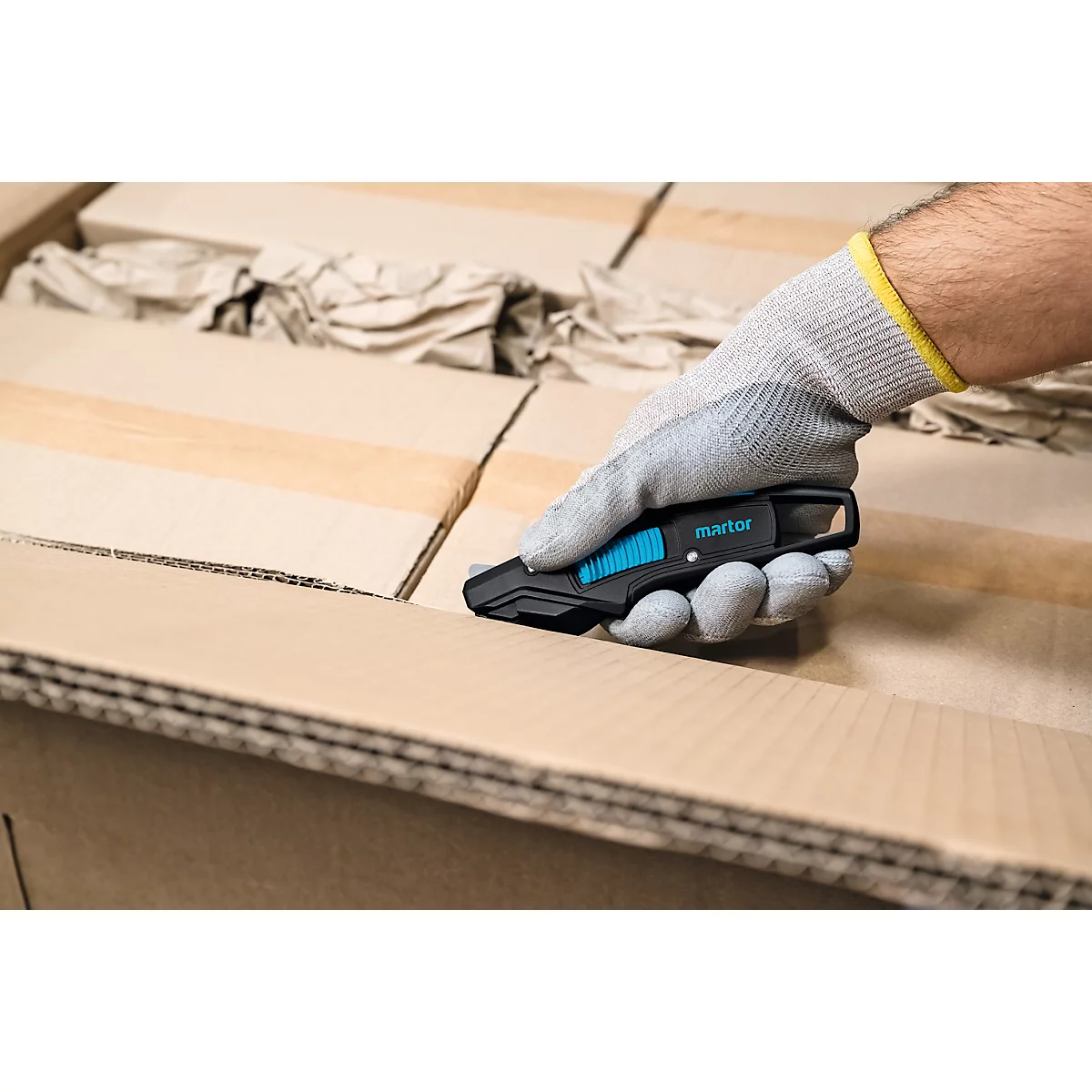 Une main gantée découpe un carton avec un couteau de sécurité noir Martor.