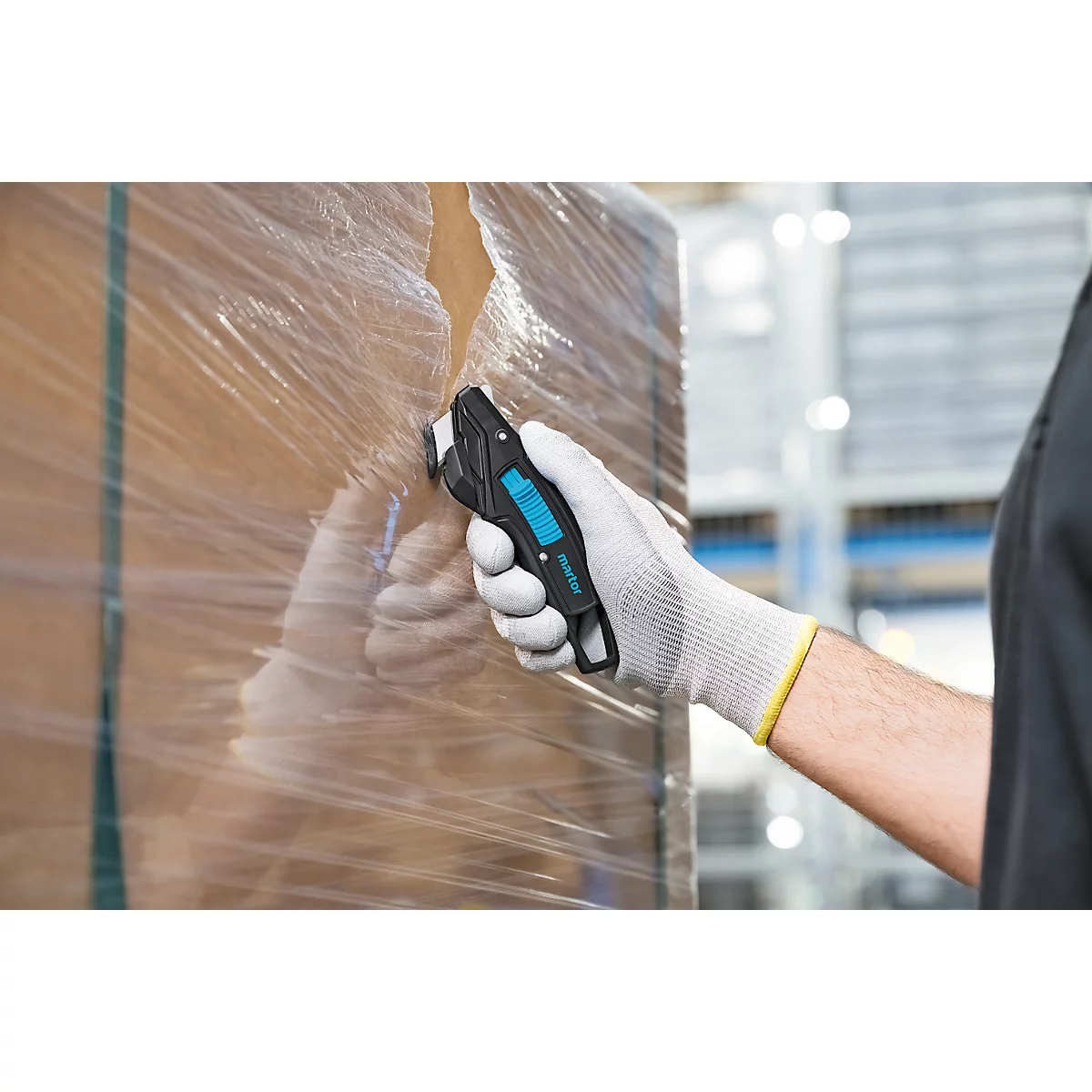 Une personne portant des gants de travail coupe le film plastique d'un colis avec un couteau de sécurité noir.