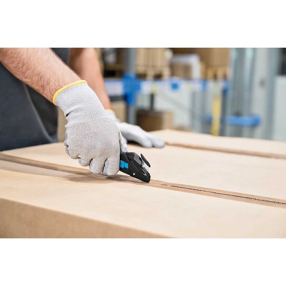 Une personne portant des gants de travail gris ouvre un carton avec un couteau de sécurité noir.