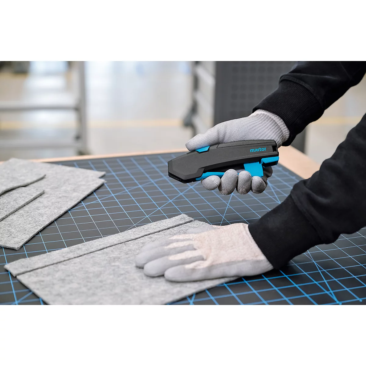 Une personne portant des gants de travail gris tient un couteau de sécurité Martor noir au-dessus d'un tapis de découpe.