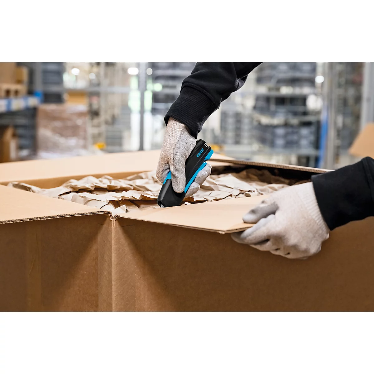 Une personne avec des gants ouvre un carton avec un couteau de sécurité bleu et noir.