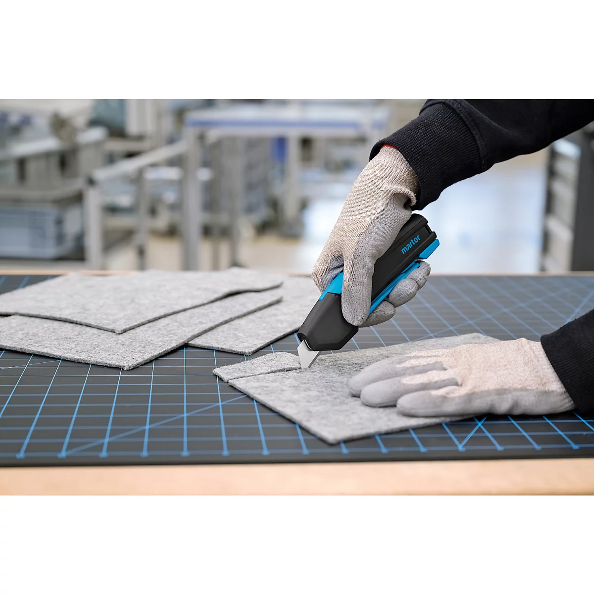 Une personne portant des gants de travail découpe un morceau de feutre gris avec un couteau Martor sur un tapis de découpe.