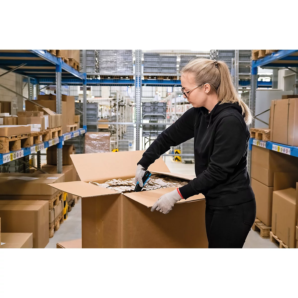 Une femme en tenue de travail ouvre avec concentration un carton d'expédition dans un entrepôt entre des rayonnages à palettes.