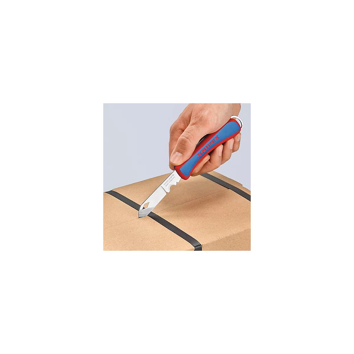 Un couteau avec une poignée bleue et rouge coupe une boîte. L'inscription KNIPEX est clairement visible.