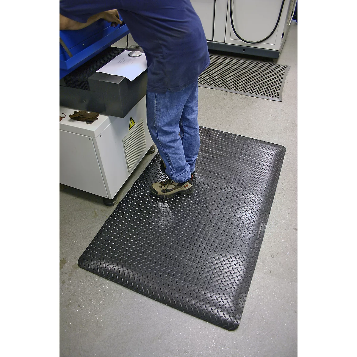 A man stands on a black, patterned workplace mat. In the background, a machine and gray floor.