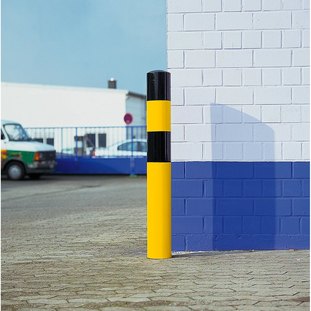Yellow and black bollard in front of white and blue brickwork. Vehicles and a gate in the background.