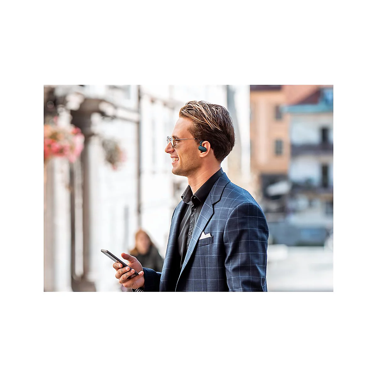 Ein Mann mit Brille und KopfhÃ¶rern lÃ¤chelt, wÃ¤hrend er ein Smartphone hÃ¤lt. Er trÃ¤gt einen Anzug, im Hintergrund eine Stadt.