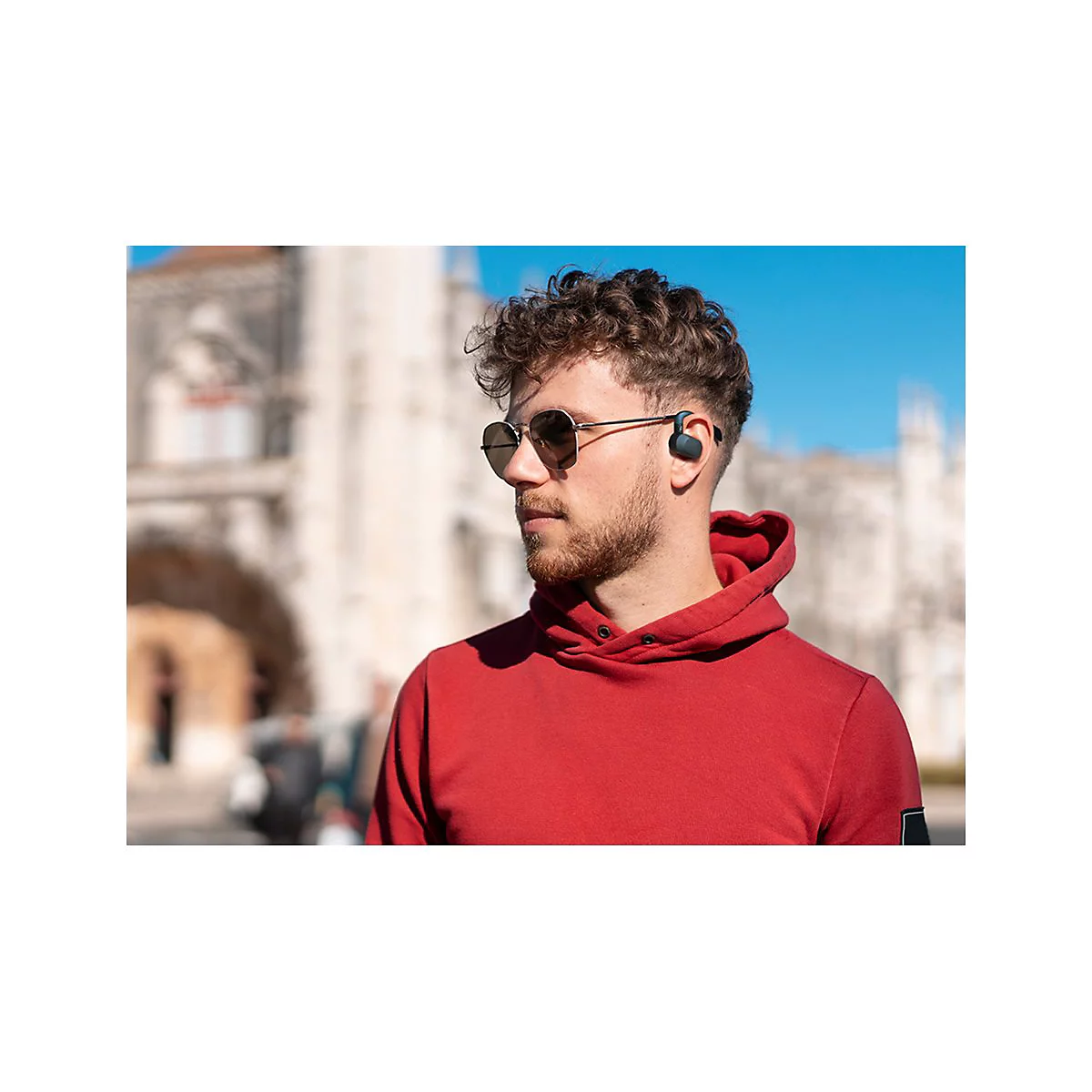 Mann mit Sonnenbrille und KopfhÃ¶rern, roter Hoodie. Im Hintergrund GebÃ¤ude und blauer Himmel.