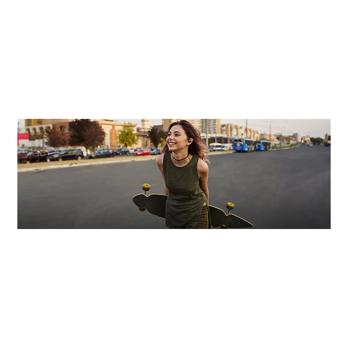 Junge Frau mit Longboard lÃ¤chelt auf der StraÃŸe. Sie trÃ¤gt KopfhÃ¶rer, ein grÃ¼nes Top und eine Halskette.