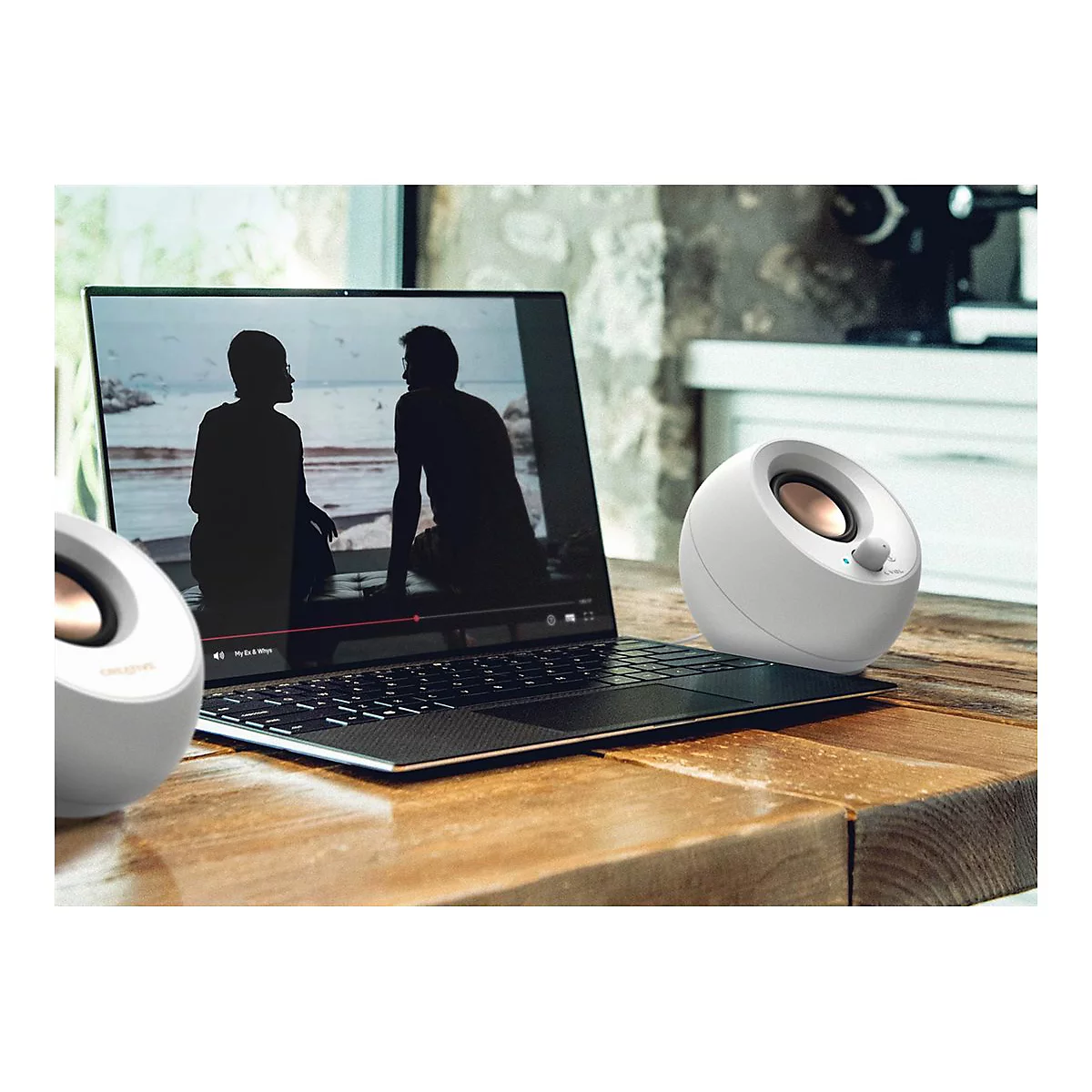 Zwei weiße Lautsprecher neben einem Laptop, auf dem ein Video abgespielt wird. Auf einem Holztisch platziert.